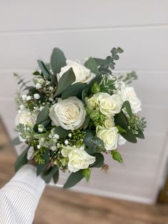 a bridal bouquet with white roses and greenery is held by someone's hand