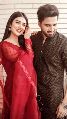 two people standing next to each other in front of a brick wall and wearing red sari