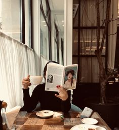 a woman sitting at a table reading a book and holding a coffee cup in front of her face