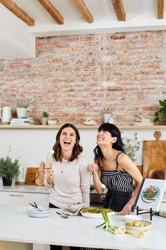 two women laughing while preparing food in a brick walled kitchen with white counter tops and exposed beams