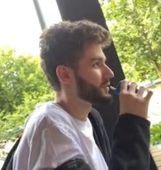 a man with a beard and white shirt is brushing his teeth while sitting on a bench