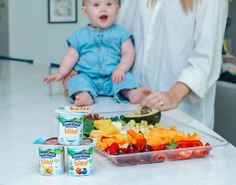 a woman holding a baby in front of a tray of fruit and yogurt