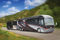 a silver and black bus driving down a road next to a green mountain covered with trees