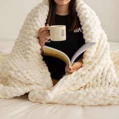 a woman is holding a book and drinking coffee while wrapped in a blanket on her bed