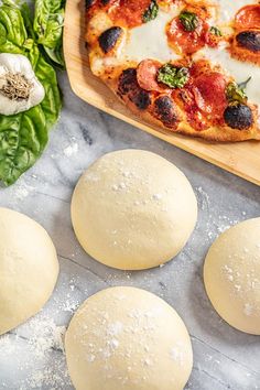 four pizza doughs sitting on top of a wooden cutting board next to basil leaves