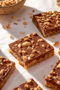 several pieces of chocolate peanut butter bars on top of a white table with bowls of nuts