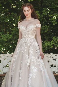 a woman in a wedding dress standing next to a bench with trees and bushes behind her