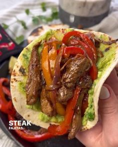 someone is holding up a taco with meat and peppers on it in front of a plate