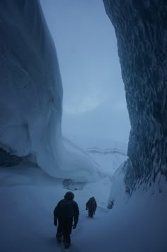 two people are walking through the snow in front of an ice cave on a cloudy day