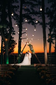 a bride and groom standing in front of the sunset at their outdoor wedding ceremony with string lights