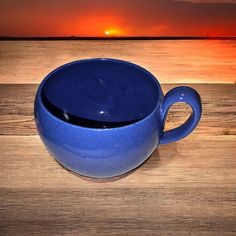 a blue cup sitting on top of a wooden table next to the ocean at sunset