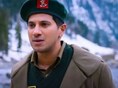 a man wearing a green hat and jacket in front of some snow covered mountains is looking at the camera