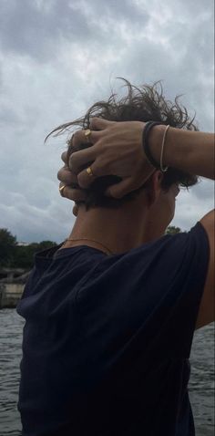 a man with his hands on his head standing in front of the water and cloudy sky