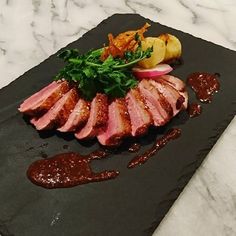 some meat and vegetables are on a black stone plate with sauces, potatoes and greens