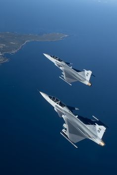 two fighter jets flying side by side in the sky above water and land area near an island