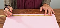 a woman measuring fabric with a tape measure on a wooden table next to a pink piece of cloth