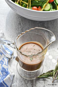 a glass pitcher filled with dressing next to a bowl of vegetables on a marble table