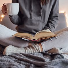 a woman is sitting on her bed while reading a book and holding a coffee cup