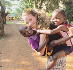 a woman holding two children in her arms on a dirt road with trees and bushes behind her