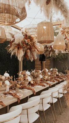 a long table set up with white chairs and hanging lights over it, surrounded by feathers