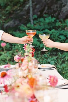 two people holding wine glasses over a table with flowers on it and greenery in the background