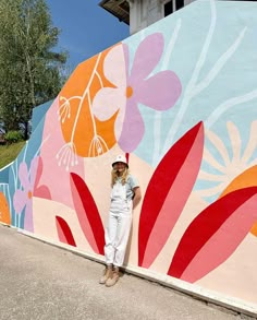 a woman standing in front of a wall with flowers painted on it and wearing a hat