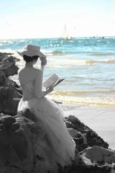 a woman sitting on rocks reading a book by the ocean with a sailboat in the background