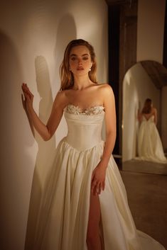 a woman in a white dress standing next to a wall and posing for the camera