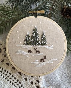 a cross stitch christmas ornament hanging on a tree branch with pine trees in the background