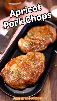 two pork chops in a black pan on a wooden table next to a knife and fork