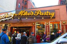 a group of people standing in front of a pastry shop on a city street with cars parked nearby