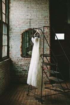 a wedding dress hanging on a rail in front of a brick wall and stairs with a mirror