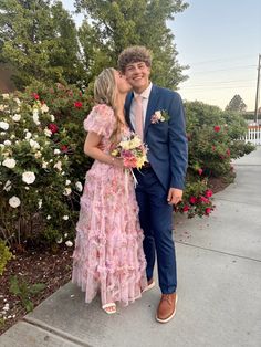 a man and woman are kissing on the sidewalk in front of some bushes with flowers