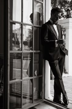 a man in a tuxedo leaning against a door and looking at his cell phone