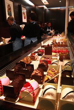 many different types of desserts on display in a buffet area with people standing around