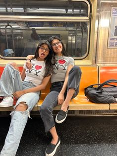 two young women sitting on a bench in front of a subway car with their arms around each other