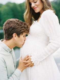 a pregnant couple standing next to each other in front of a body of water and trees