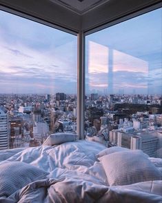 a bed in front of a large window overlooking a cityscape at sunset or dawn