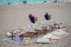 a table set up on the beach with purple flowers and place settings in front of it