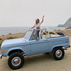 a woman standing on top of a blue truck with a dog in the back seat