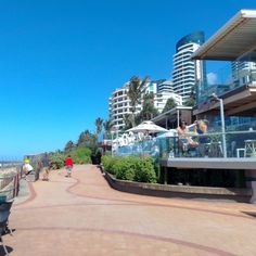 people are walking on the sidewalk near some buildings by the water and beach with umbrellas