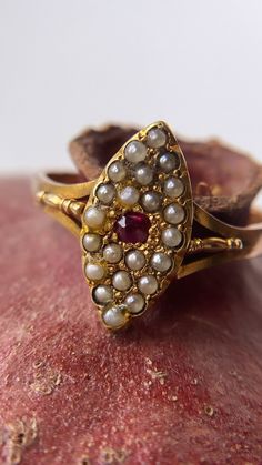 A beautiful antique Victorian navette ring featuring a natural ruby surrounded by beautiful seed pearls. The ruby takes centre stage - a pigeon red pop of colour that has such a presence for its size! It is bright, strong and powerful! Surrounding the ruby are 21 creamy grey, seed pearls, all original, in a beautiful navette form. What I particularly like is the sweet gold detailing in between each seed pearl and the patina is so wonderful, an old matt yellow gold. With beautiful detailed split Vintage Marquise Ruby Ring Gift, Vintage Marquise Ruby Ring As Gift, Vintage Marquise Ruby Ring For Gift, Vintage Marquise Ruby Ring In Red, Vintage Marquise Ruby Wedding Ring, Shaena Targaryen, Ruby And Pearl Ring, Navette Ring, Antique Ruby Ring