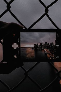 a person holding up a cell phone in front of a fence with the city skyline behind it