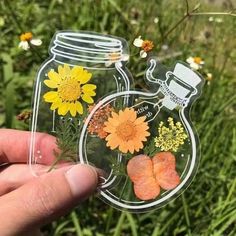 a hand holding up a glass jar with flowers in it on top of some grass