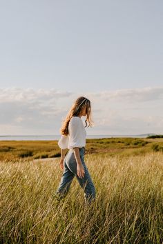 a woman walking through tall grass in the middle of an open field on a sunny day