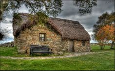 an old stone house with a thatched roof and bench in the grass near trees