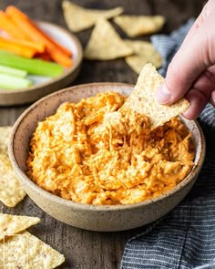 a hand dipping a tortilla chip into a bowl of dip with carrots and celery