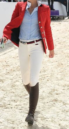 a woman walking across a sandy field wearing a red jacket and white riding breeks