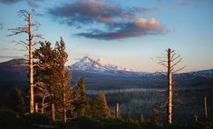 the sun is setting on a mountain range with trees in front of it and snow - capped mountains in the distance