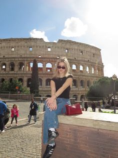 a woman sitting on top of a brick wall in front of an old building with people walking around